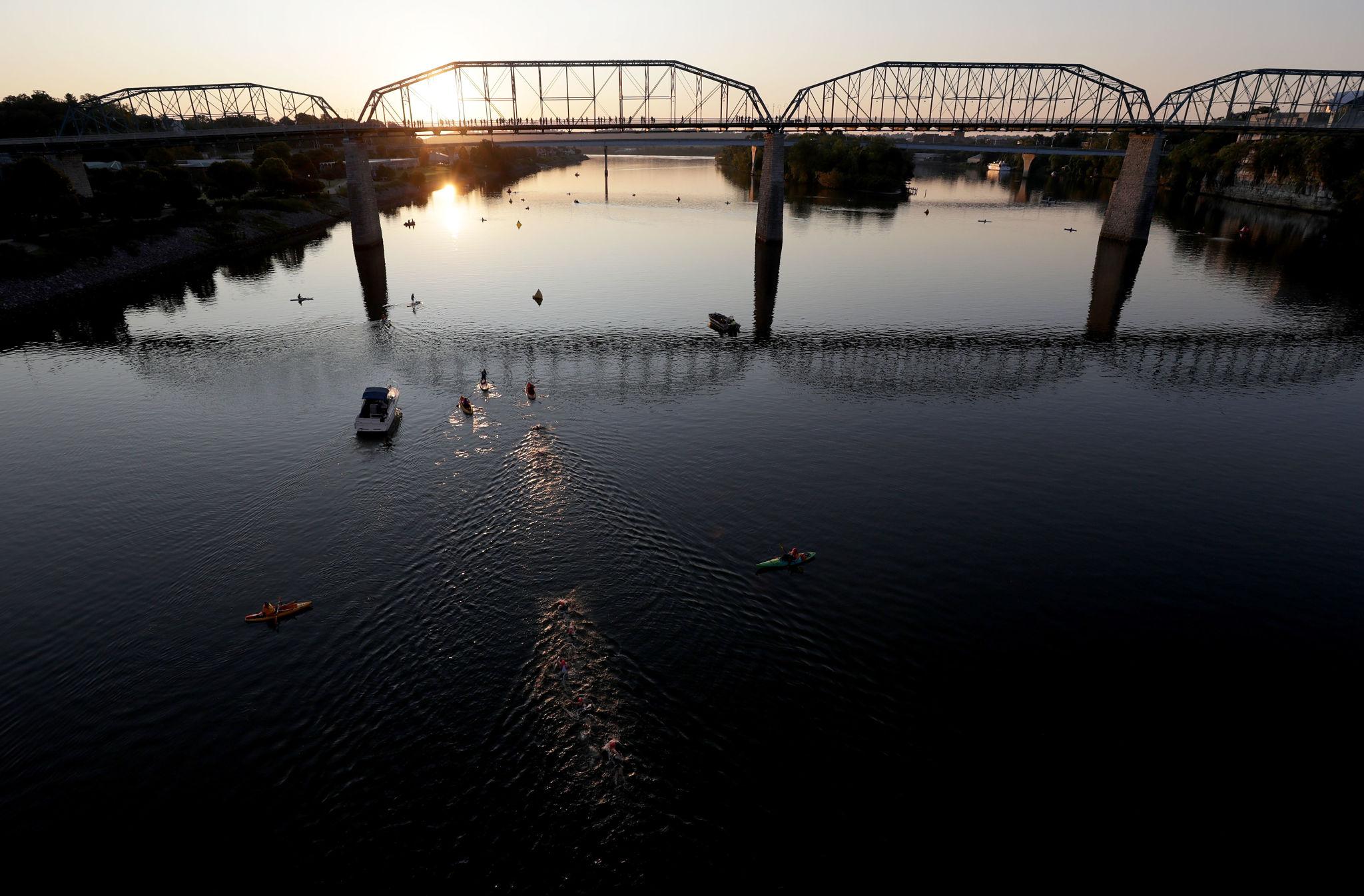 The Walnut Street Bridge over the Tennessee River at sunrise, with open-water swimmers, kayak safety escorts, and support boats on the IRONMAN 70.3 Chattanooga swim course